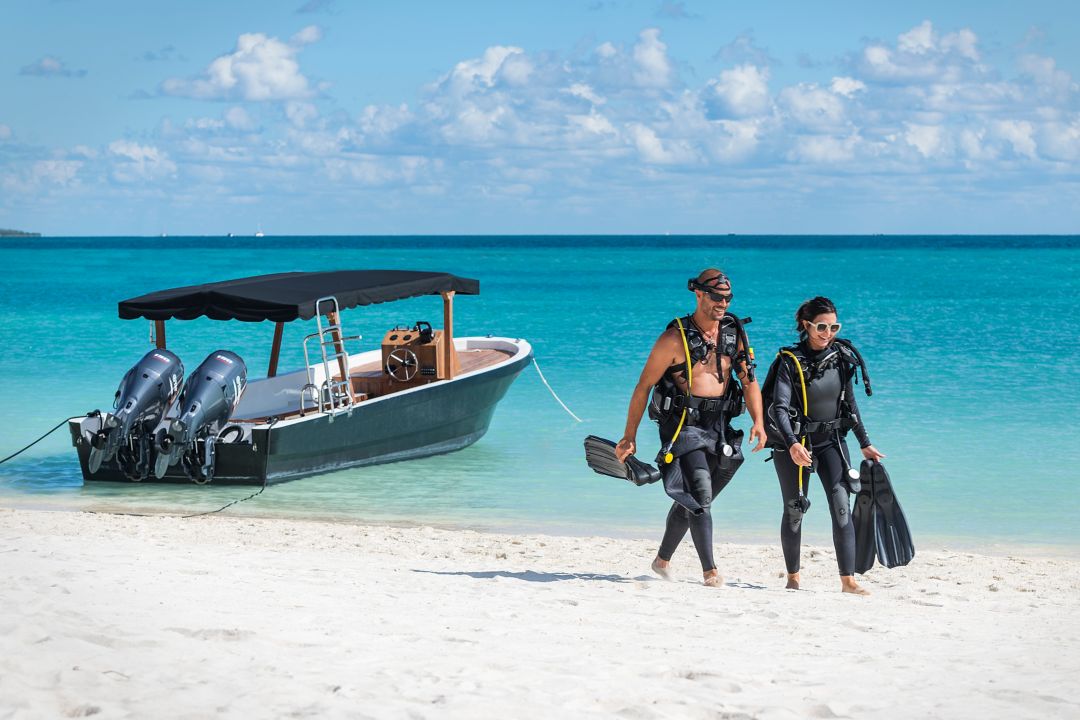 Woman and man wearing wet suits and scuba gear walk along the beach while small motor boat floats anchored at the shore.